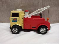 Side view of the vintage M&M Tow Truck toy showing yellow cab, red rear tow body, and white tow arm with hook.