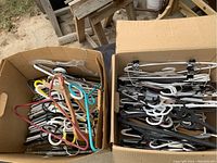 Two open cardboard boxes filled with assorted plastic, wooden, and metal clothing hangers of various colors and styles