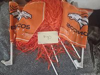 Two Denver Broncos orange auto flags with white plastic clips and four orange pompoms on a grey textured fabric surface