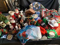 Wide angle image showing a variety of Christmas linens, candles, Santa and snowman figurines, nativity set, and other holiday decorations arranged on a black tablecloth for sale.