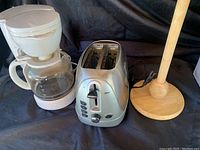 Full view of the white drip coffee maker, stainless steel toaster, and wooden paper towel holder grouped together on a dark cloth background.