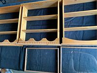 Three wooden decorative plate display shelves shown side by side, with visible curved slots for plates and multiple horizontal slats. Shelves are light-colored wood on a dark cloth background.