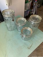 Four clear glass jar containers with metal latch-lock lids on a light blue surface. Jars vary in size and show some staining.