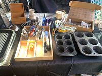 Wide shot of the baking and kitchen tools lot showing pans, measuring cups, graters, teapot, and various utensils on black tablecloth
