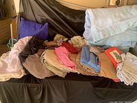 Various linens including sheets and blankets laid out showing different colors and textures including a blue blanket, purple pillow, brown and beige linens and rags.