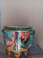 Front view of the Mexican pottery pot showing colorful hand-painted floral and geometric patterns with green textured side handles and glossy finish.