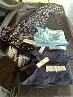 Photo showing four clothing items including black floral print dress, black dress with adjustable straps, light blue sleeveless top, and navy dress or top, all size 3X with tags.