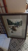Full view of framed winter mountain scene picture standing against wall on patterned carpet.