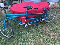 Side view showing entire blue tandem bicycle frame with front and rear handlebars, pedals, and seatpost with front seat attached, missing rear seat.