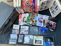 Top-down photo showing DVDs, assorted CDs, cassette tapes in cases, and VHS tapes arranged on a black cloth.
