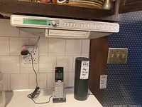 Sony wall-mounted radio/CD player with digital clock and control buttons shown mounted under a kitchen cabinet. Below it are an AT&T cordless phone handset with charging base and an Amazon Alexa speaker.