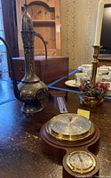 Engraved metal samovar tall with handle and spout, brass candle holder with faux red roses and white candle, and two small round instruments alongside a wooden base.