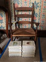 Wooden child's chair with rush seat in front of floral wallpaper, set atop carpet, with books stacked on floor in front of chair.