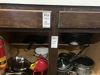 Photo showing several pots and pans with lids, a large glass bowl, and a red plastic colander inside a wooden cabinet space.