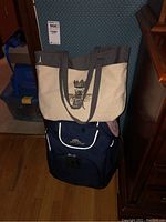 Front view of beige canvas shoulder bag with black bee wearing crown design placed on top of navy blue High Sierra carry-on backpack bag with wheels