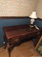 Wooden vanity table by Dixie shown from an angle, with a ceramic lamp on top and the table pushed against a wallpapered corner wall. Visible surface scratches and marks on the table top. Two drawers with brass handles visible on the front.
