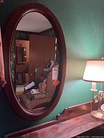 Oval mirror resting against green wallpapered wall, reflecting interior room details with visible wooden frame and lamp nearby.