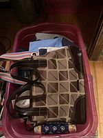 Photo of a beige and brown pocketbook with a geometric pattern, brown leather corners, and multicolored striped handles placed in a red bin with other items.