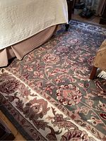 Photo shows part of the rug under a bed and near a chair, highlighting the floral pattern and tan border, with visible wear on edges.