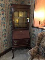 Full view of wooden secretary desk with glass-paned upper cabinet and two lower drawers, siting in corner against wallpapered walls.