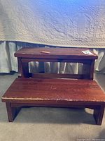 Photo of a reddish wooden two-step stool with scratches and wear on the surface and edges.