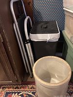 Two folding step stools leaning against a wall next to a black trash basket with lid and a large ceramic canister in front on a patterned rug.