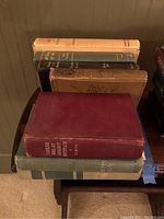 Nine vintage books stacked on a wooden surface showing worn aged covers and varied colors.