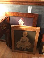 Two framed portraits stacked on floor; one depicts a young girl with a basket and the other a girl with long braided hair.