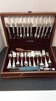 Wooden hinged case open with two tiers showing 12 knives in top tier and assorted flatware and serving utensils below.