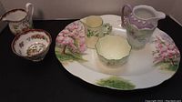 Photo showing multiple ceramic items including a large oval Royal Albert plate with hand-painted cherry blossom trees, a yellow creamer with floral handle, and a scalloped edge cream bowl with green trim.