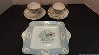 Two double handled cups with floral patterns and gold trim on matching floral saucers behind a square plate featuring a bird on a branch with a blue and floral border on a gold trimmed plate.
