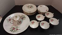 Full set laid out showing 6 side plates, 1 serving plate, 6 saucers, 5 cups, and sugar bowl with lid. All have pink rose floral pattern and gold trim edges.