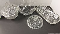 Four cut crystal serveware pieces on black surface: covered bowl with lid, three-footed bowl, extra lid, rectangular plate