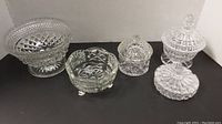 Four clear cut glass pieces: two covered candy dishes, one three-legged bowl, and one pedestal bowl arranged on black background.