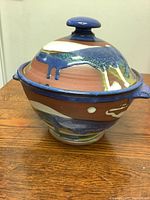 Side view of handmade pottery bowl with lid, showing multi-colored glaze and artistic drip details, lid knob and handles on bowl visible.