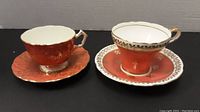 Two Aynsley tea cup and saucer sets side by side on a black surface, showing the difference between a corset-shaped cup and a standard round cup.