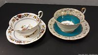 Two Aynsley tea cup and saucer sets displayed side-by-side, one floral pattern and one blue and gold