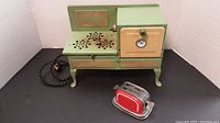 Green metal toy stove with cream-colored oven door and vented stovetop, fabric-covered cord visible.