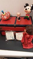 Metal toy chest with stickers and vintage Mickey Mouse pull toy, pig figurine, and red boxing glove puppet in front, with two Peanuts toys in McDonald's packaging also visible