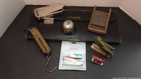 Photo showing eight vintage items including staplers, hanging scale, bell, leather vase, and a small wooden frame inside a box.