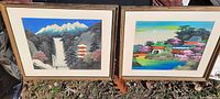 Two framed artwork pictures shown side by side on grass, depicting Asian landscapes with mountains, waterfalls, and buildings.