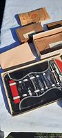 Photo showing a collection of vintage personal grooming sets arranged on a white surface, some open displaying razors and tools in cases and boxes, others still in original packaging or paper envelopes.