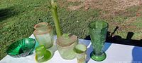 Photo showing entire collection on white surface outdoors with grass background, displaying seven distinct green and opaque glassware pieces in various shapes and sizes.