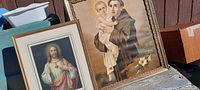 Photo shows two framed religious prints placed leaning against a wall, one with Jesus Christ and the other slightly obscured.