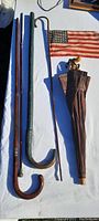 Four antique wooden canes and walking sticks with varied handles, a small early American flag attached to one, and a closed brown umbrella with wooden handle seen on a white cloth surface outdoors.