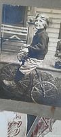 Photo of a young boy sitting on a tricycle or small motorcycle toy, wearing goggles on his head, against a porch backdrop