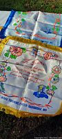 Photo of two antique silks laid flat on a white surface, one with blue tasselled edges and floral military-themed graphics, the other with gold tasselled edges and colorful military imagery and text.