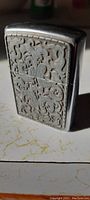 Front view of metal Zippo lighter showing detailed embossed floral or scrollwork pattern