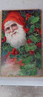 Christmas postcard showing Santa Claus with red hood and white beard surrounded by holly leaves and berries