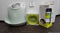 Three items shown together: BabyBjorn bib light gray, Ola Baby lime green Steam Bowl in box, bbluv stackable containers in box.